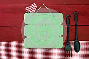 Blank rustic wood menu sign hanging by cast iron spoon and fork and red gingham tablecloth