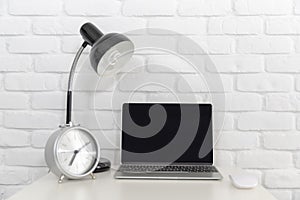 A blank screen Laptop computer on white desk with lamp and silver clock with white brick wall.