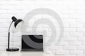 A blank screen Laptop computer on white desk with lamp and white brick wall.