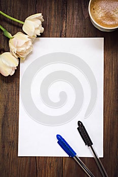 Blank sheet of paper, pens, flowers and cup of coffee on dark wooden desk.