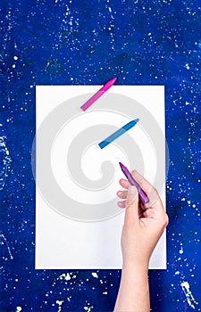 Blank sheet of white paper and hand hold crayons on galaxy-blue background