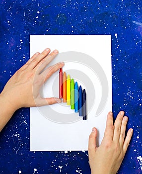 Blank sheet of white paper and hand hold multicolor crayons on galaxy-blue background