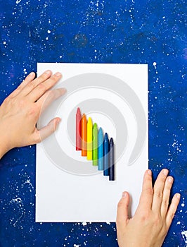 Blank sheet of white paper and hand with multicolor crayons on galaxy-blue background