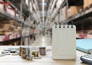 Blank note book paper stand, coin money and calculator  on wooden table with in large warehouse for writing an information.