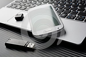 Blank Smartphone With Green Reflection Lying On Business Notebook Keyboard Next To An USB Storage, All Above A Carbon Layer