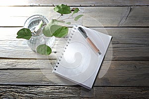 Blank spiral notepad with grid paper, pen and a glass vase with some leaves on a rustic table made of old wooden planks, mockup