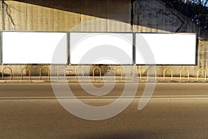 Blank street billboards on a grunge concrete wall in an underpass. Metal bollards, sidewalk and asphalt road in front.