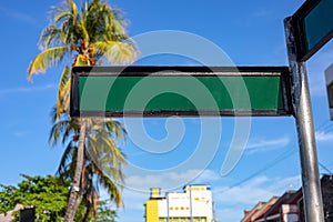 Blank street sign on sunny urban landscape with palm trees and yellow building. Green metallic sign plate on pillar