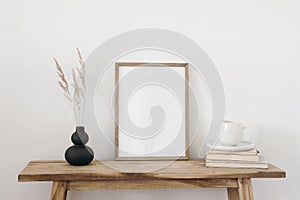 Blank vertical wooden picture frame mockup. Organic shaped black vase with dry grass on table, bench. Cup of coffee on