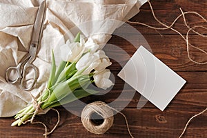 Blank wedding invitation card and bunch of spring flowers on wooden table. White tulips, textile rag, twine, romantic wedding gree