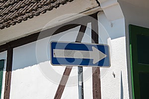 Blank white arrow on blue background street sign on pole with white wall with brown trim in background. Brown roof and green shutt