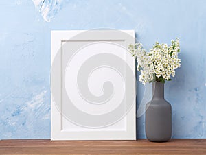 Blank white frame, flower in vaze on brown wooden table against pastel blue concrete wall with copy space. Mock up.