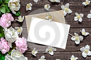 Blank white greeting card in frame made of pink roses and white jasmine flowers and envelope on rustic wooden background