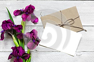 Blank white greeting card with purple iris flowers bouquet and envelope on white wooden background
