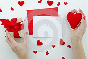 Blank white greeting card with red envelope Female`s hands hold gift box and red felt heart symbol of love. Valentines