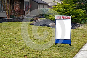 Blank white lawn sign in the grass in front of a house. A sign on top says Sale Pending.