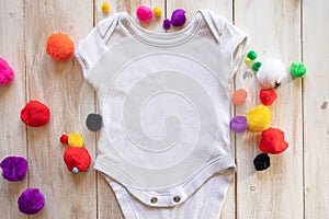 Blank, white mock up baby bodysuit shirt on a light white-washed wooden background surrounded by multi-coloured craft pom poms
