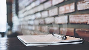 A blank white notebook and silver color pen on wooden table with brick wall