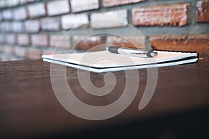 A blank white notebook and silver color pen on wooden table with brick wall