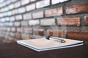 A blank white notebook and silver color pen on wooden table with brick wall