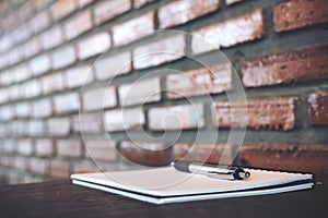 A blank white notebook and silver color pen on wooden table with brick wall