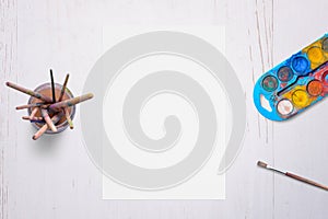 Blank white paper on desk, surrounded by brushes and watercolors