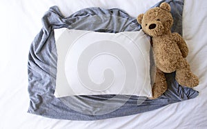 Blank white rectangle cushion laying on a grey scarf with a white duvet background and child`s teddy