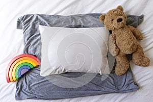 Blank white rectangle cushion laying on a grey scarf with a white duvet background and child`s teddy and rainbow