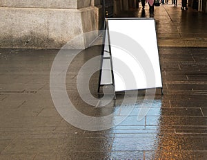 Blank white sandwich sign outside in daytime on dark wet sidewalk near a building.