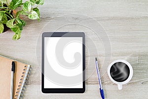 Blank white screen tablet computer with hot coffee cup, green plant, notebook and pens on wooden desk