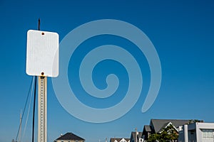 Blank white sign on a metal post against a clear cloudless blue sky. There are the tops of houses in the background