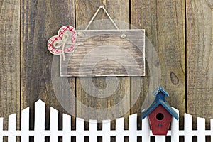 Blank wood sign with plaid heart hanging over white picket fence with birdhouse