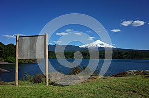 Blank wooden information board with view of snow capped Villarrica volcano and lake Villarrica.