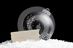 Blank wooden sign and black and silver glittering ornament sitting in snow