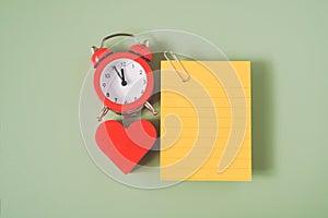 Blank yellow note paper with paper clip, red heart shape and analog alarm clock on green background
