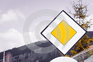 Blank yellow road sign meaning ` Yield pedestrians ` and `Main high  priority road` on sky background in nature, forest mountains.