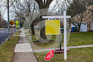 Blank yellow sign on a white post on a lawn in the front yard of a house near a sidewalk and a street.