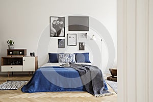 Blanket on blue bed with pillows in retro bedroom interior with radio on cabinet and posters. Real photo