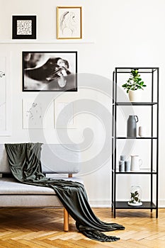 Blanket on grey sofa next to shelves in simple white living room interior with posters. Real photo