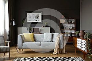 Blanket and pillows on grey settee in retro living room interior with armchair and poster. Real photo