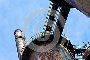 Blast furnaces and smokestack ringed with walkways as part of an abandoned old industrial steel mill site, viewed from below, blue