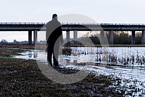 A bleak, moody, winter edit of a figure standing next to a lake, looking out to a motorway bridge, out of focus in the background