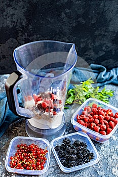 Blender with oatmeal, plastic plates, containers with raspberry, blackberry, red currant, cherry on a stone background