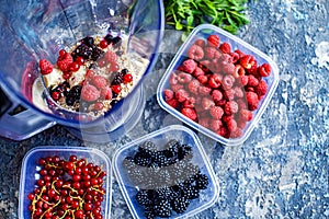 Blender with oatmeal, plastic plates, containers with raspberry, blackberry, red currant, cherry on a stone background