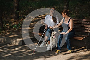 Blind caucasian woman sitting on bench with guide dog and pregnant friend.