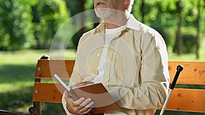 Blind man reading book in Braille sitting on bench in park, social adaptation