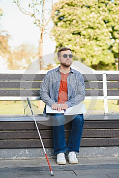 Blind man reading braille book, sitting on bench in summer park, resting