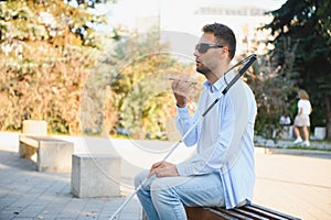 Blind man with a walking stick sits on a bench uses a smartphone on the background of a city.