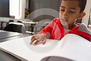 Blind schoolboy reading a braille book at desk in a classroom