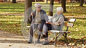 Blind senior man with sunglasses on sitting with his wife on a bench in the park
